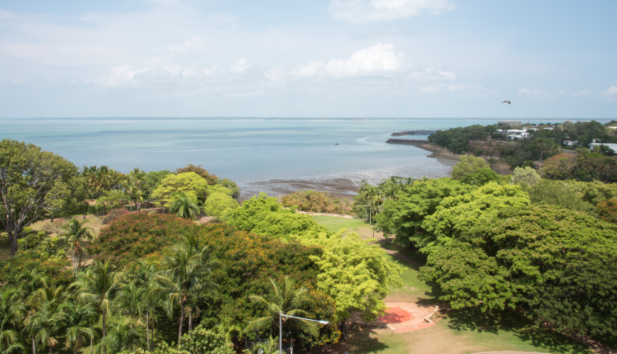 A scenic view of lush green trees and a coastal shoreline under a partly cloudy sky, with calm blue water stretching into the distance and a few buildings visible among the trees.