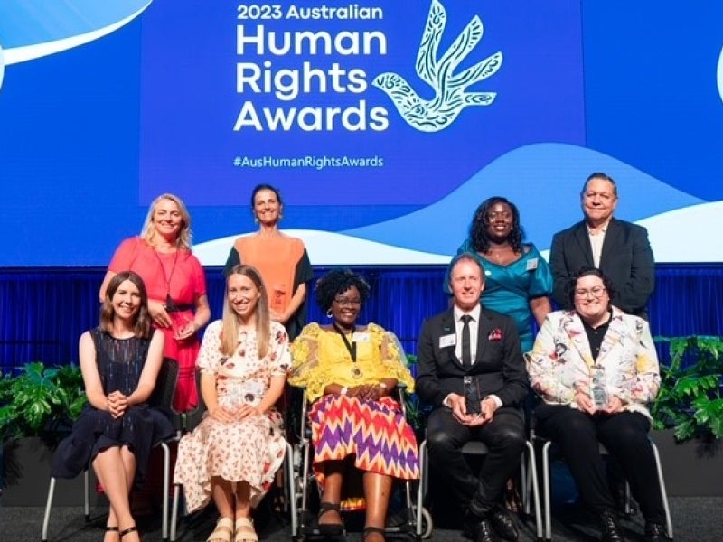 Ten people pose and smile on stage in front of a blue screen displaying “2023 Australian Human Rights Awards"