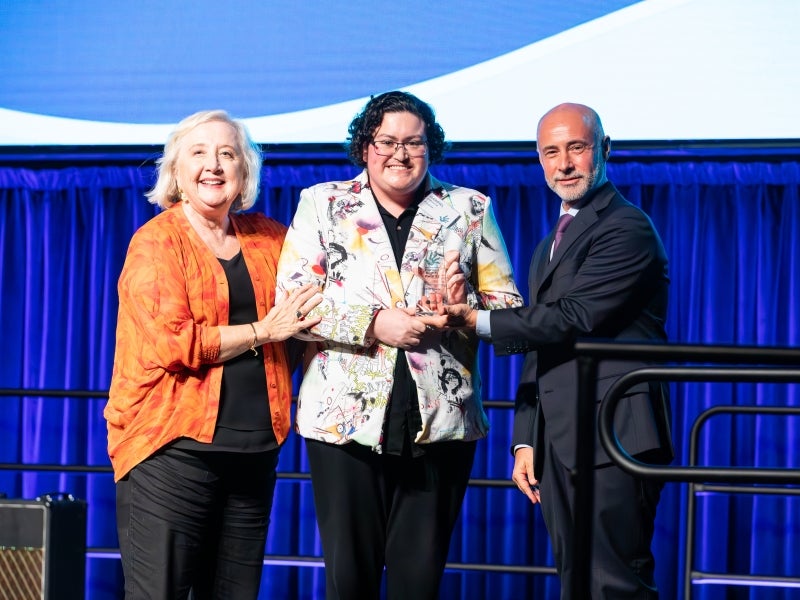 Three people stand on a stage with blue curtains, smiling at the camera. The person in the center holds a glass award, while the people on either side touch the award in celebration.