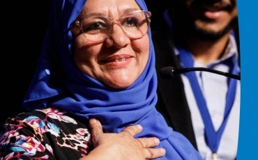 Woman in a blue headscarf and floral top stands before a podium with her hand on her chest to accept an award while in the background a man looks on beaming. 