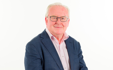 An older man with short gray hair and glasses, wearing a blue blazer over a light pink collared shirt, smiles at the camera against a plain white background.