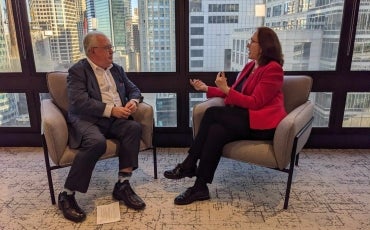 Two people in business attire sit facing each other in armchairs by large windows with a city skyline in the background, engaged in conversation. A sheet of paper lies on the floor near one person.