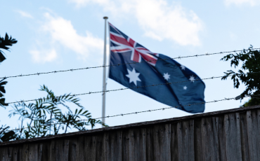 A flapping Australian flag seen through the barbed wire of a high stone fenc